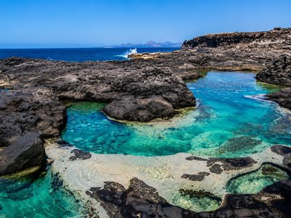 Los Charcones Lanzarote — wild natural swimming pools cut into black volcanic rock on the southwest coast near the Pechiguera lighthouse, with crystal-clear turquoise water and the Atlantic beyond