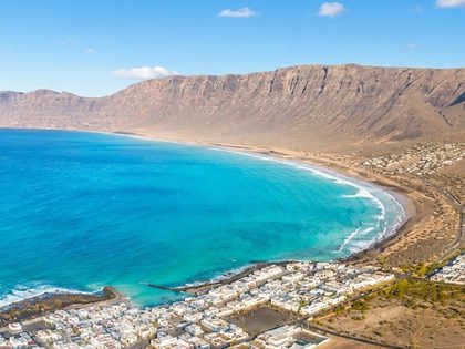 Aerial view of Caleta de Famara Lanzarote — long curved golden-sand beach, whitewashed village, turquoise Atlantic surf, and the volcanic Risco de Famara cliffs in the background