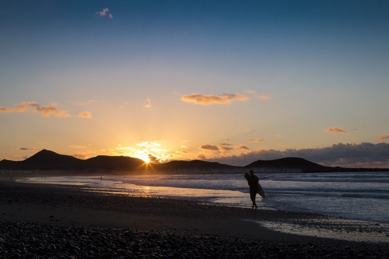 Famara beach Lanzarote at sunset — surfer walking on pebbly shore with volcanic cliffs silhouetted against golden sky