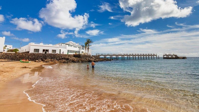 Northern Lanzarote coast — Arrieta village shoreline with whitewashed buildings, volcanic coastline and Atlantic Ocean under a bright blue sky