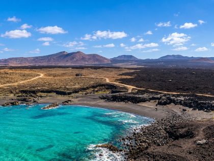 Playa de las Malvas Tinajo Lanzarote — wild cove of dark volcanic sand and gravel with the eroded ancient lava flows of Timanfaya in the background and turquoise Atlantic water