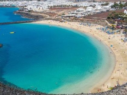Aerial view of Playa Dorada in Playa Blanca Lanzarote — sheltered golden-sand bay with turquoise calm water, black lava breakwater, sun loungers and the Montana Roja volcano in the background