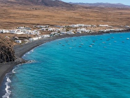 Playa Quemada Lanzarote — small cove of coarse black volcanic sand in a sheltered semicircular bay, whitewashed fishing village with boats anchored in the turquoise water